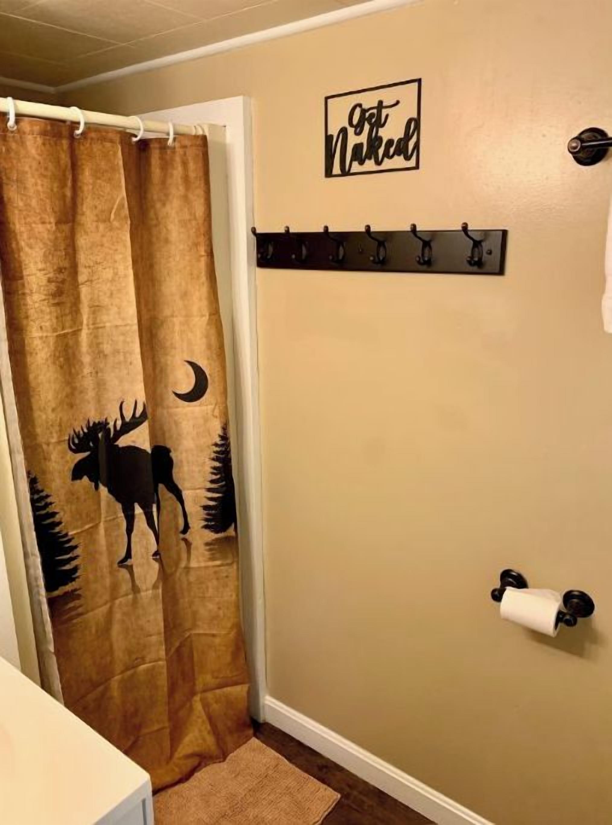 A bathroom with a brown shower curtain featuring a moose and crescent moon design, a towel rack with a 'First Naked' sign, and a toilet paper holder.