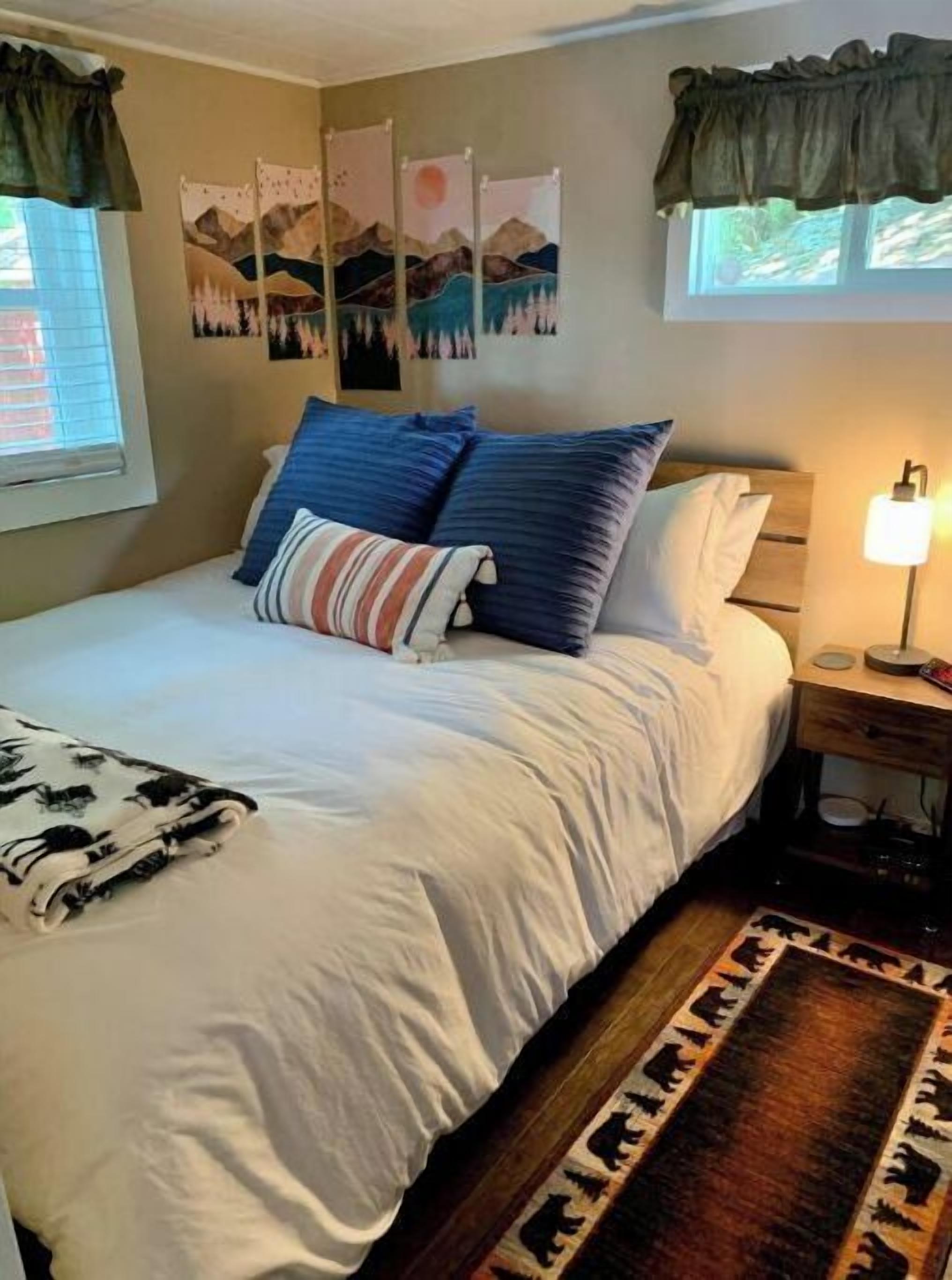 A neatly made bed with white bedding and multiple pillows, a wooden nightstand with a lamp, and a patterned rug on the floor.