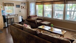 Una sala de estar acogedora con un sofá, sillones y una mesita de centro. Se ve una ventana con persianas, un dispensador de agua y una pequeña mesa con una planta.