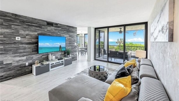 A modern living room with a flat-screen TV, grey sofa, and a balcony with a view of the ocean. - Luxury condo on the beach  (Naples)