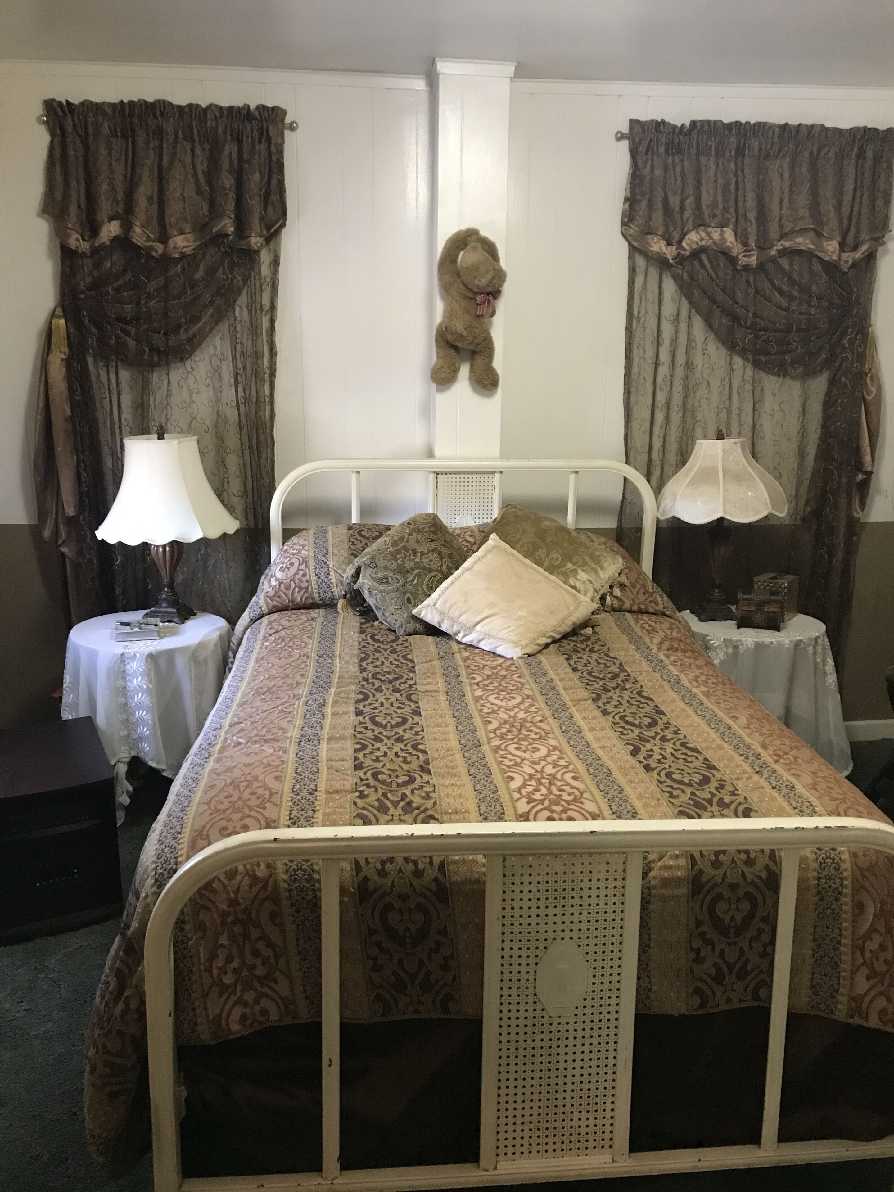 A single-bed room with a metal frame, patterned bedding, two bedside tables with lamps, a teddy bear on the wall, and curtains.