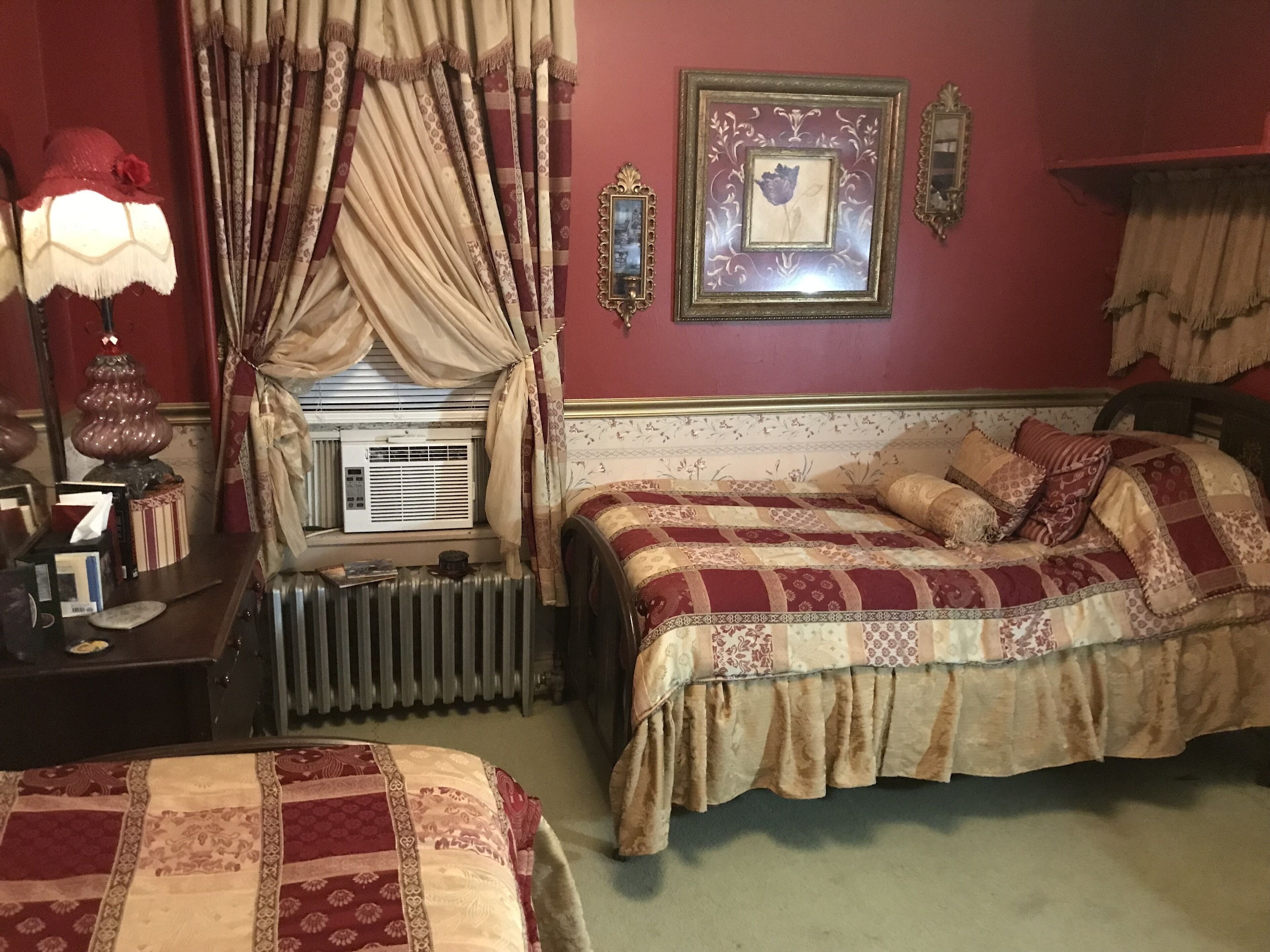 A bedroom with two beds, a radiator, a window with curtains, a lamp, and framed pictures on the wall.