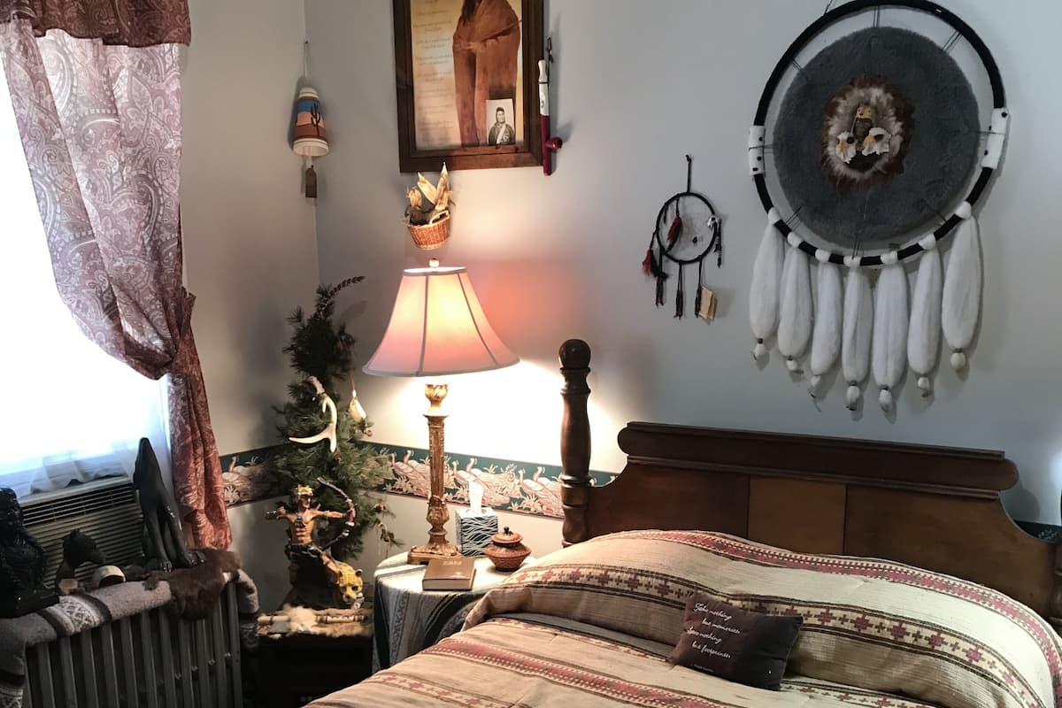 A bedroom with a large bed, a nightstand with a lamp, a framed picture on the wall, and a dreamcatcher.