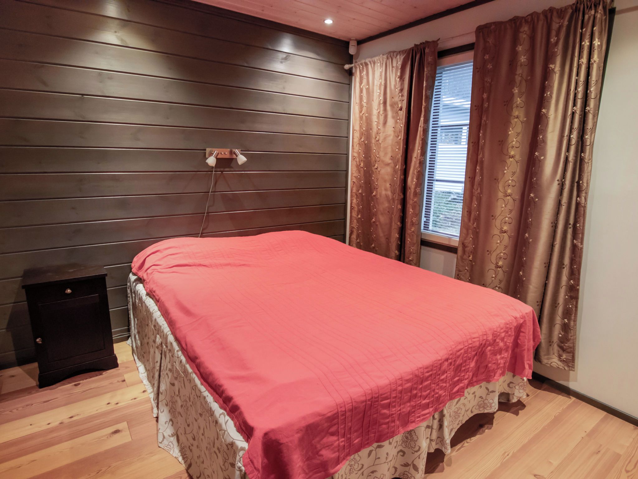 A bedroom with a wooden headboard, a bed with a red bedspread, a nightstand, a window with curtains, and a ceiling light.