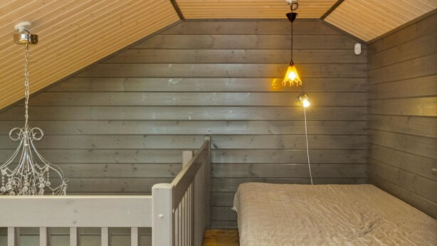 A small, cozy room with wooden walls and a sloped ceiling. It features a single bed with a quilted cover, a wooden railing, and a hanging light fixture. - Pulkka 3 by Interhome (Salla)