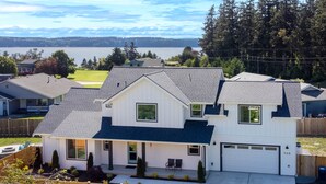 A two-story house with a garage, a covered porch, and a view of a lake. - 5BR vacation home Hot Tub/Game Room/Water Views/BBQ/firepit/LG Yard (Camano)
