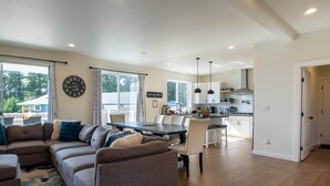 A modern living room with a sectional sofa, dining table, and kitchen area with white cabinets and pendant lighting. - 5BR vacation home Hot Tub/Game Room/Water Views/BBQ/firepit/LG Yard (Camano)