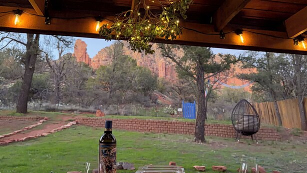 An outdoor dining area with a wooden table, plates of food, a bottle of wine, and a view of a natural landscape with trees and rocks. - Red rock views ♦ Private access to the forestry ♦ The perfect romantic escape (Sedona)