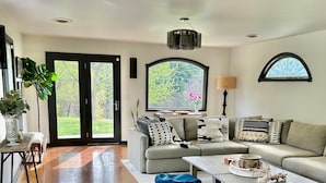 A modern living room with a grey sofa, a coffee table, and blue ottomans. There is a large window with a view of greenery outside. - Dog-Friendly Cottage w/ Creek, Barn, Game Room, Fire Pit, Hot Tub, Sleeps 10 (Hillsville)