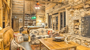 A cozy log cabin interior with a wooden dining table, plush seating, a bookshelf, and a stone fireplace. - River Access, Mtn Views! All-Season Cullowhee Base (Cullowhee)