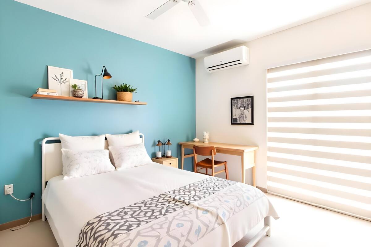 Quarto com cama, escrivaninha e cadeira. Há uma estante com livros e uma planta.