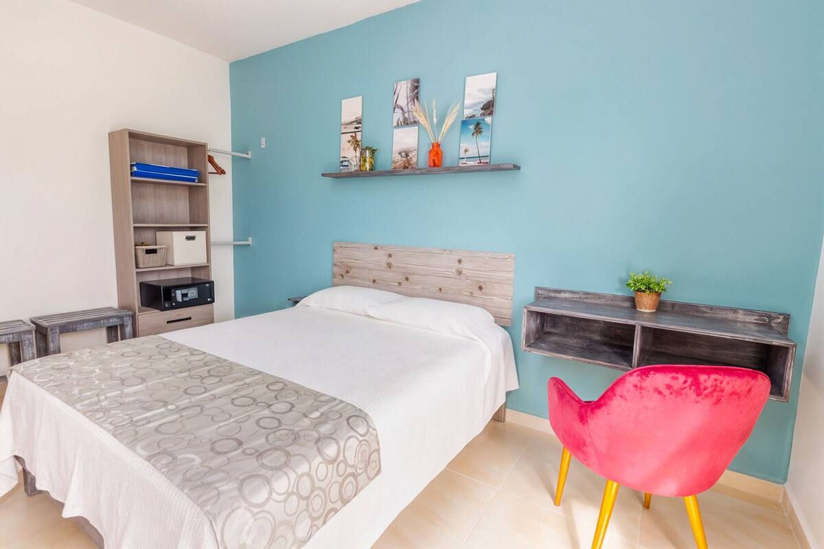 Quarto com cama, cabeceira de madeira, prateleira com obras de arte, cadeira vermelha e escrivaninha com computador.