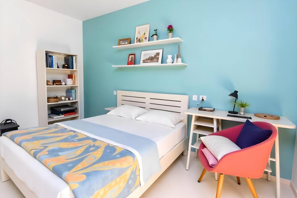 Quarto com cama, escrivaninha, cadeira e estantes com livros e objetos decorativos.