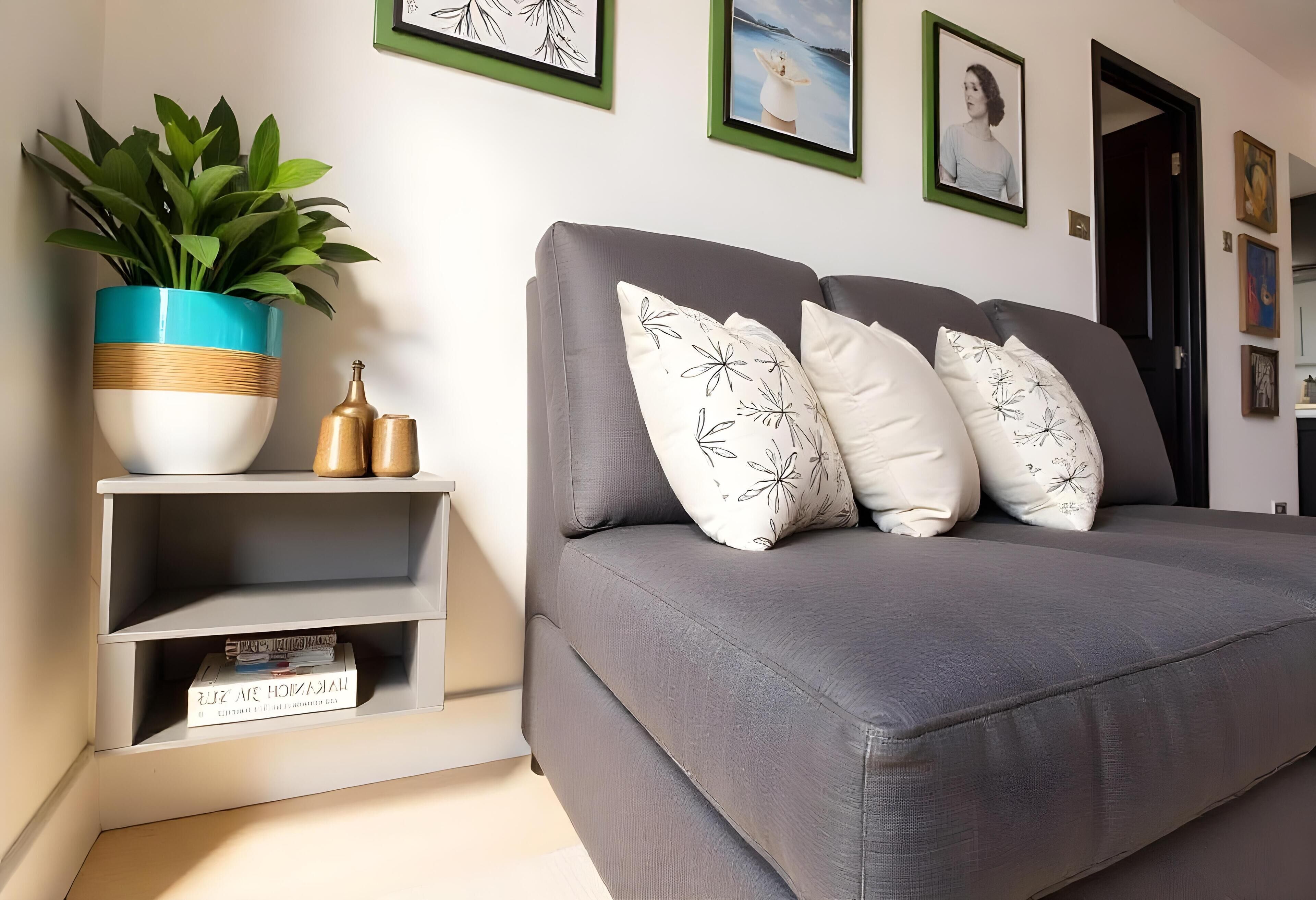 Een moderne woonkamer met een grijze bank, zwart-wit gestreepte kussens, een blauwgroene plant met witte bloemen in een houten pot en een witte plank met boeken en decoratieve objecten.