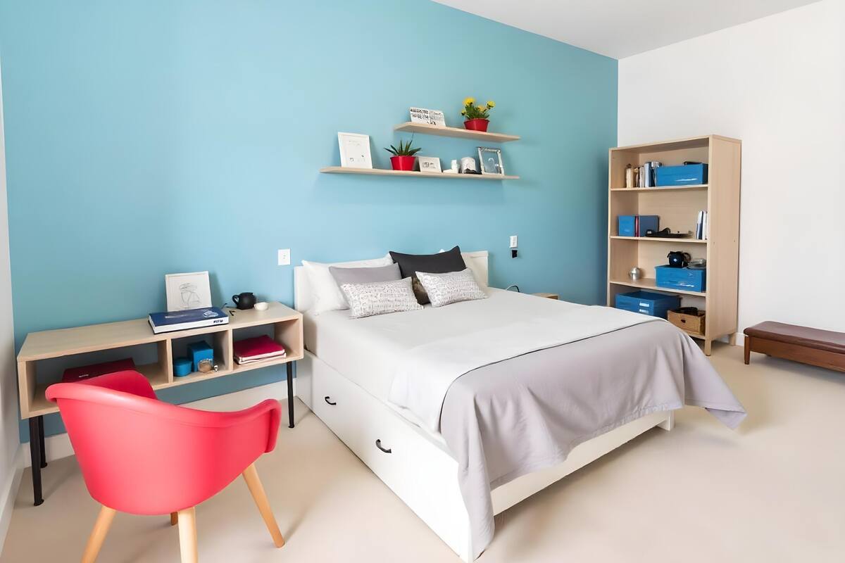 Quarto com cama, escrivaninha, cadeira, estantes e prateleiras.