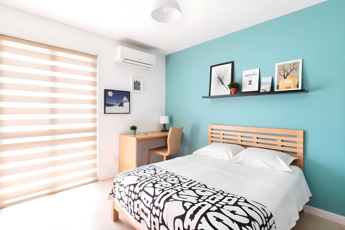 Quarto com cama, escrivaninha e cadeira. Há uma janela com persianas, ar-condicionado fixado na parede e quadros emoldurados.