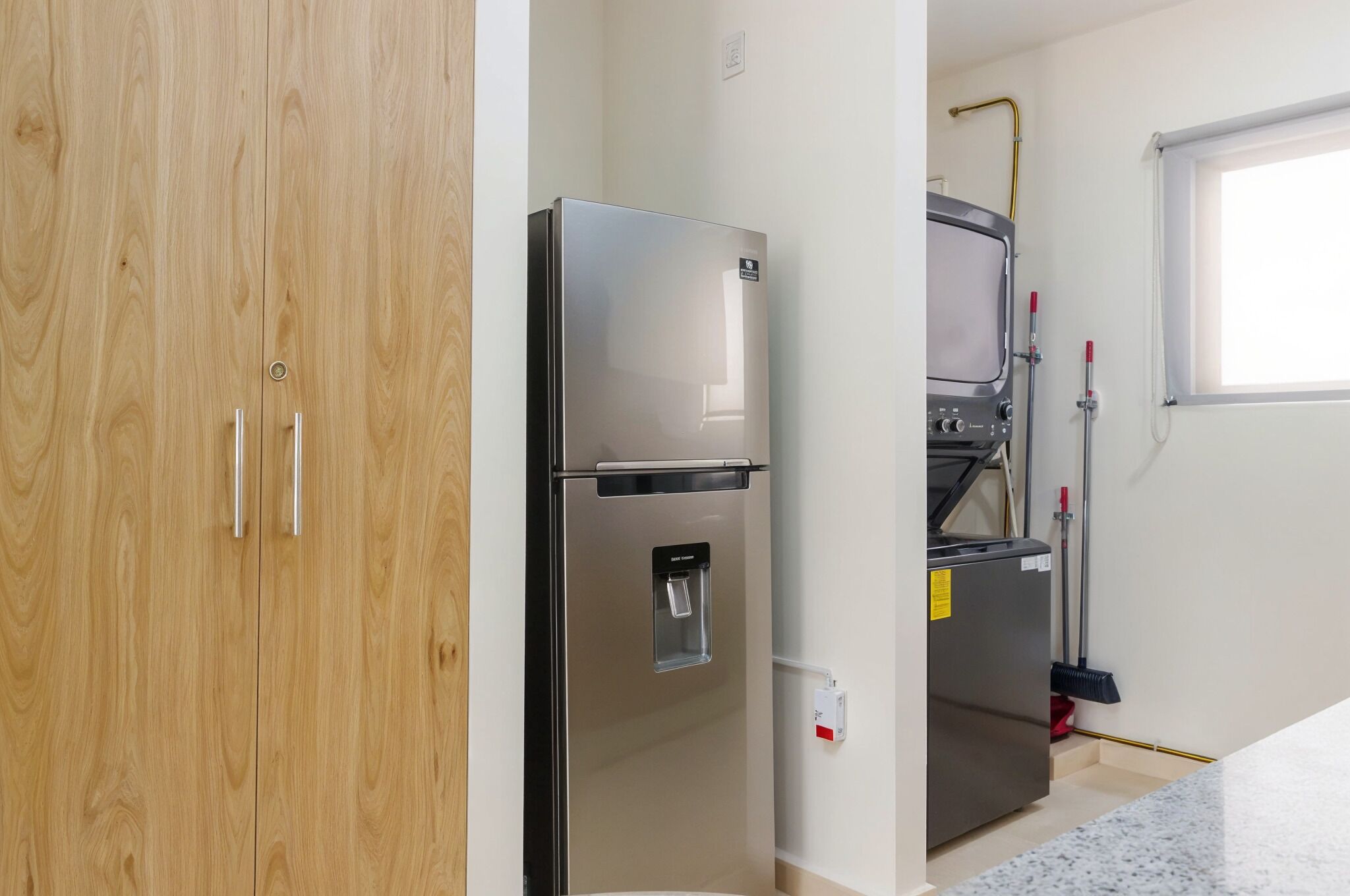 Una cocina con un refrigerador de acero inoxidable, un microondas y un dispensador de agua. Hay una escoba apoyada en la pared y una ventana con vista al exterior.