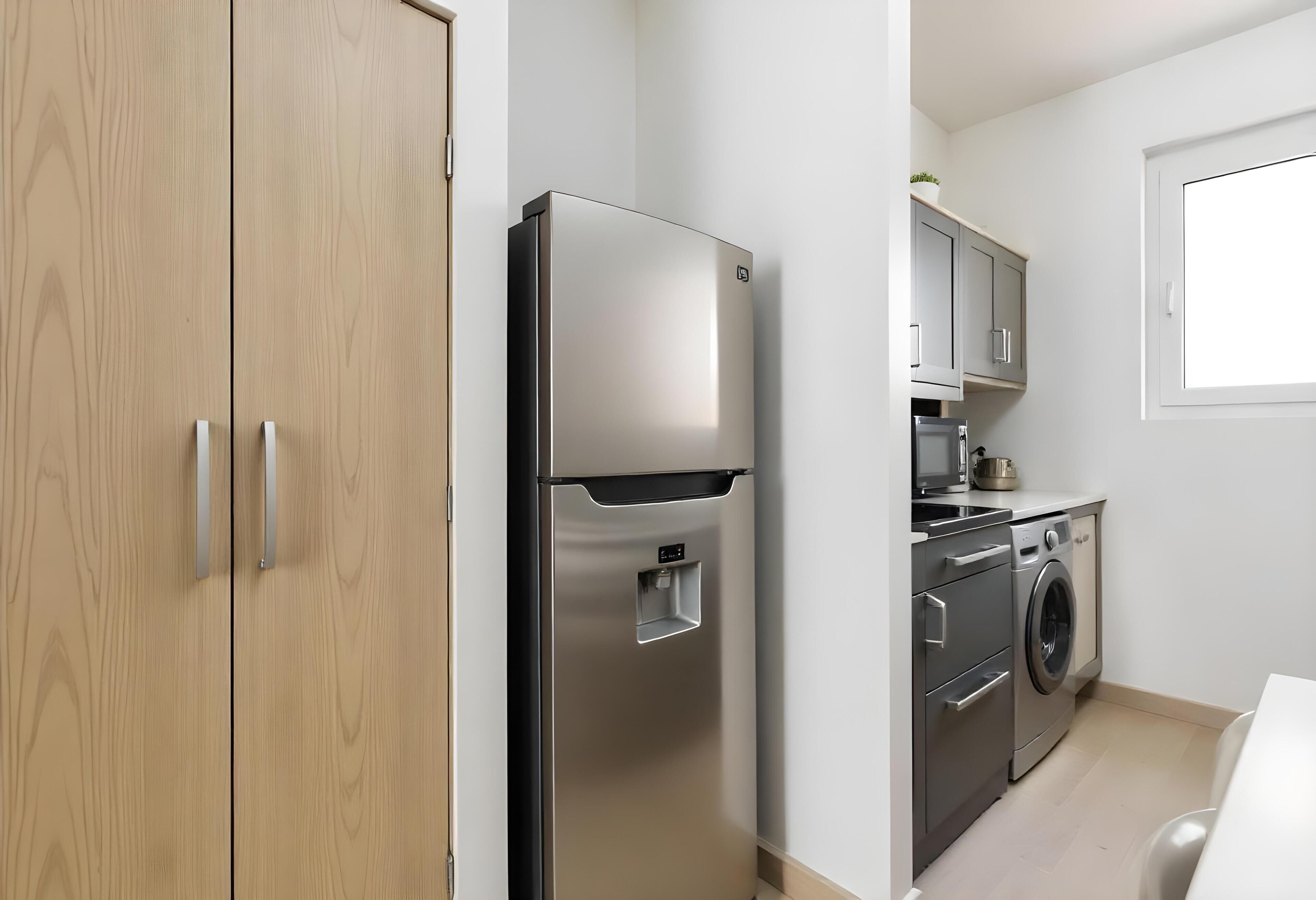 A modern kitchen with a stainless steel refrigerator, a built-in washing machine, and wooden cabinets.