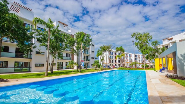 A swimming pool with blue circular steps, surrounded by modern buildings with balconies and palm trees. - Bright & Relaxing Full Apartment (Cancun)