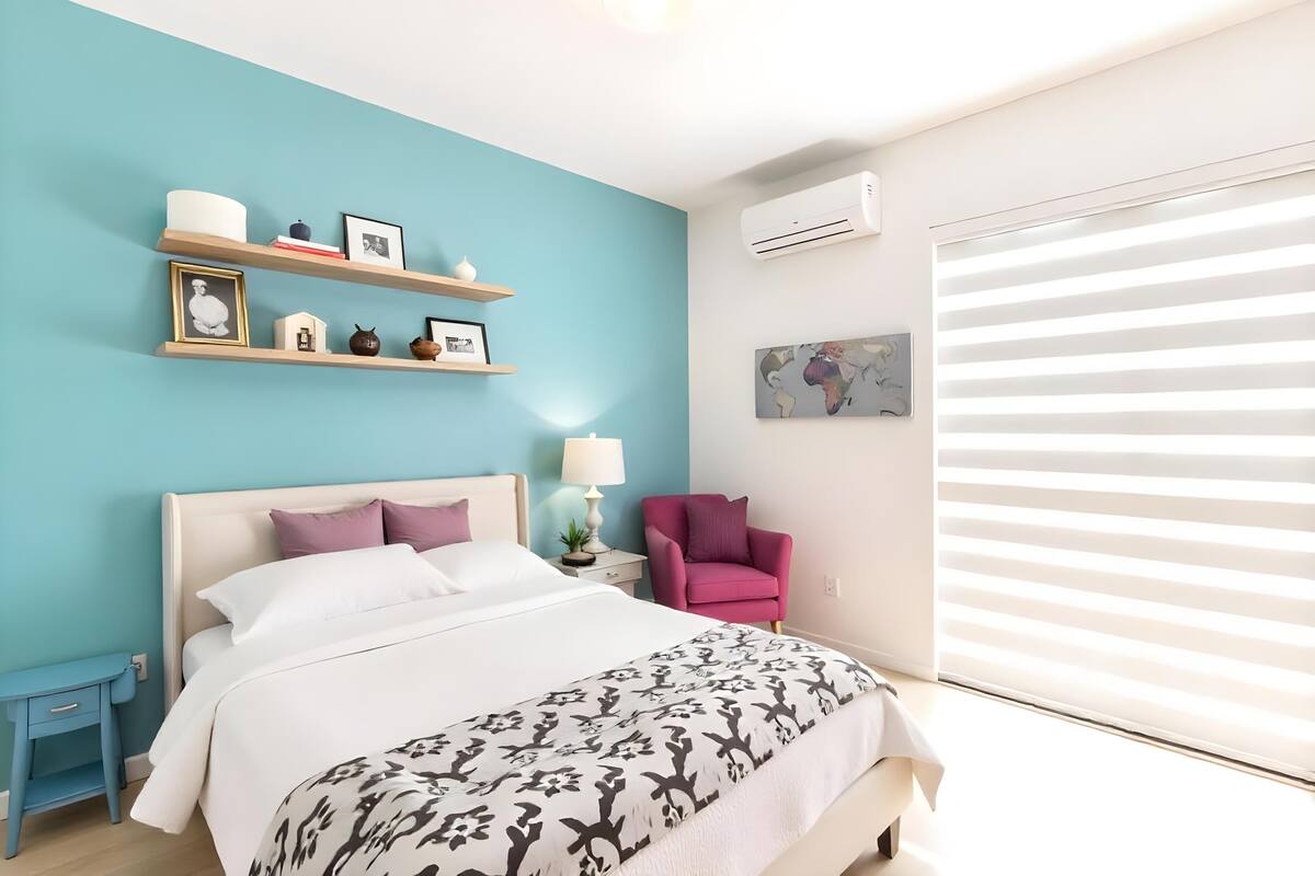 Quarto com cama, mesa de cabeceira, cadeira, prateleira com objetos decorativos e janela com persianas.