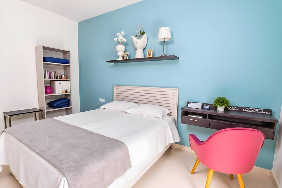 Quarto com cama, escrivaninha, cadeira e prateleiras com livros e objetos decorativos.