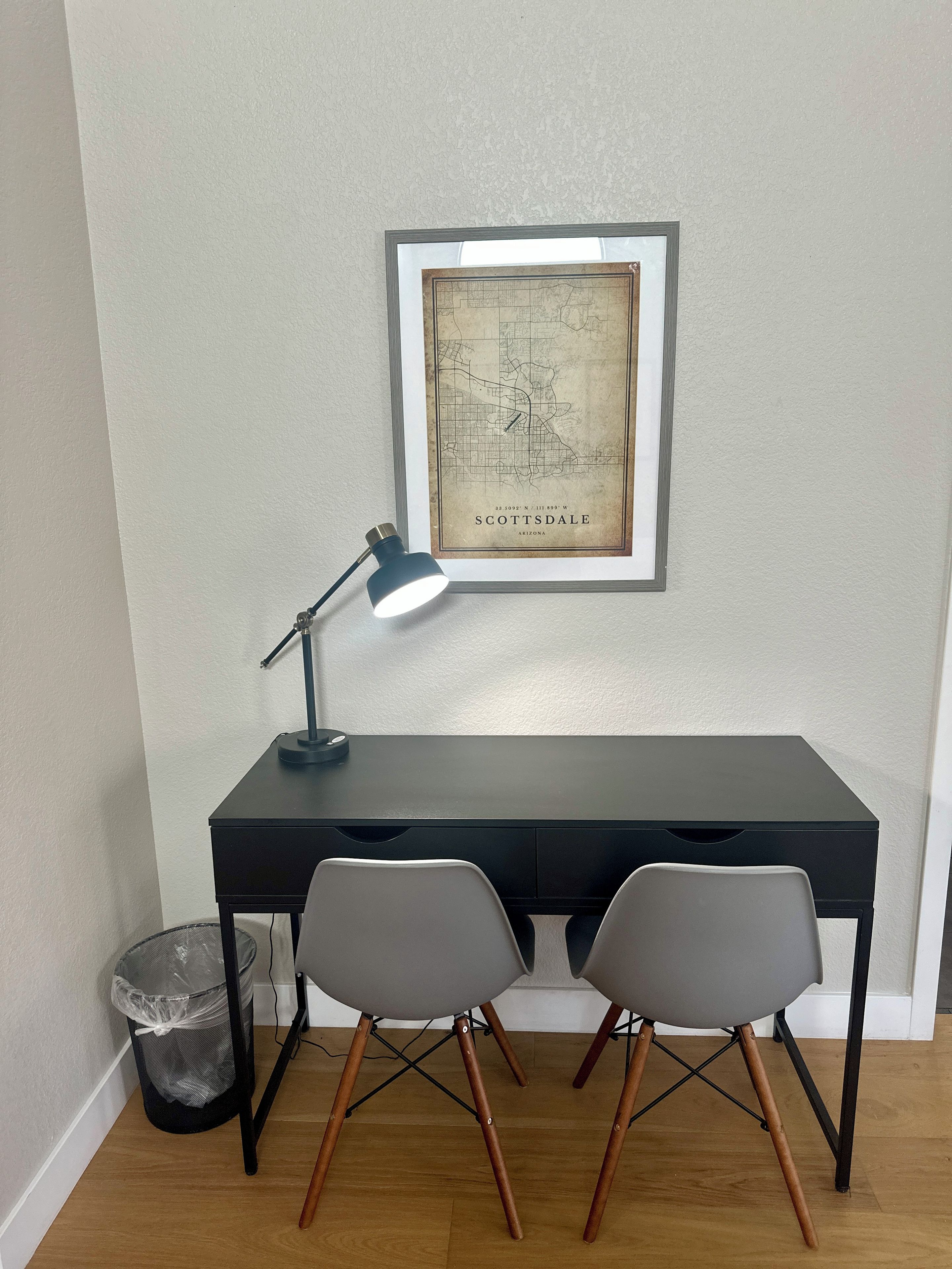 A desk with two chairs, a framed map on the wall, and a desk lamp.