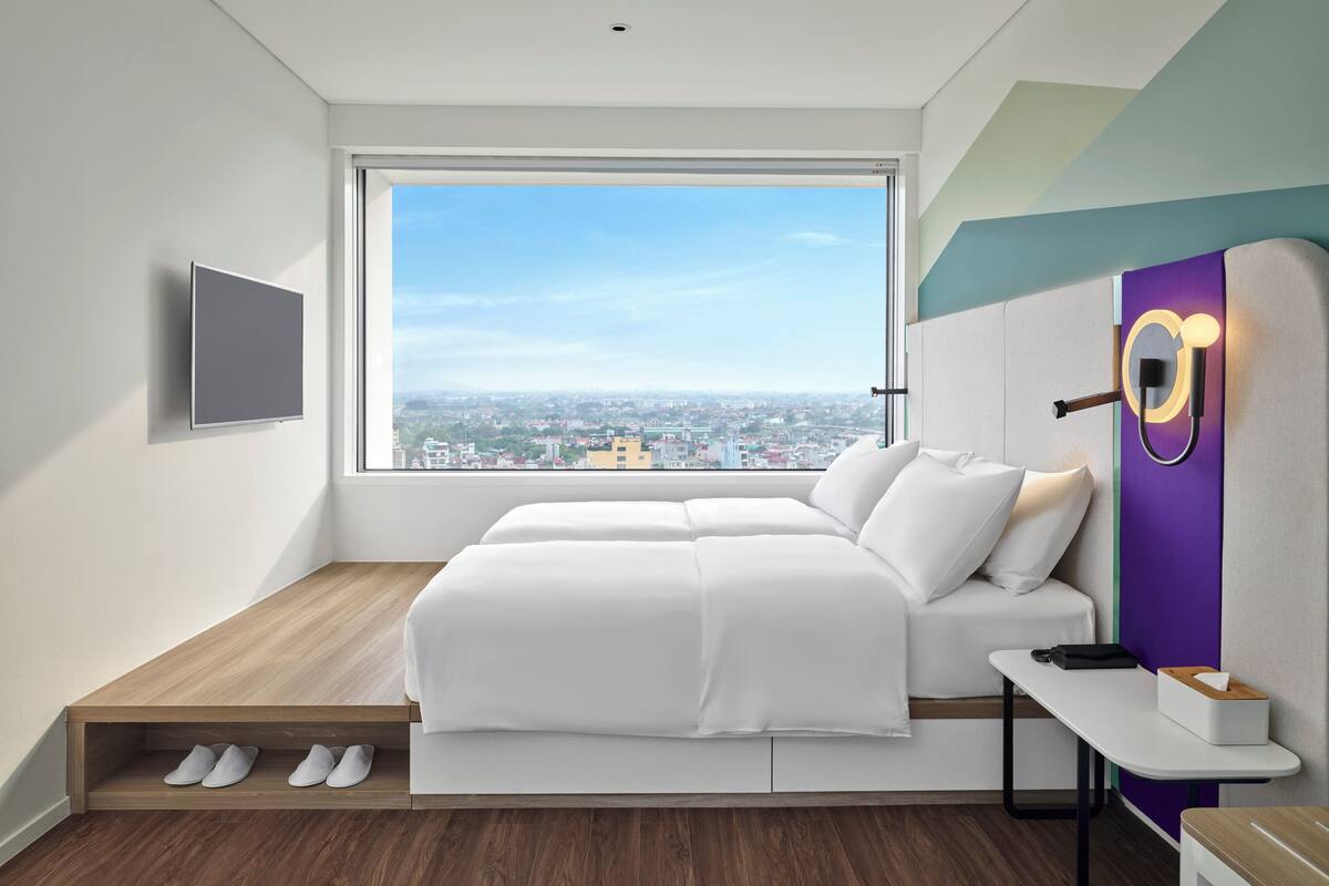 A modern bedroom with a large window, a bed, a desk, and a wall-mounted TV.