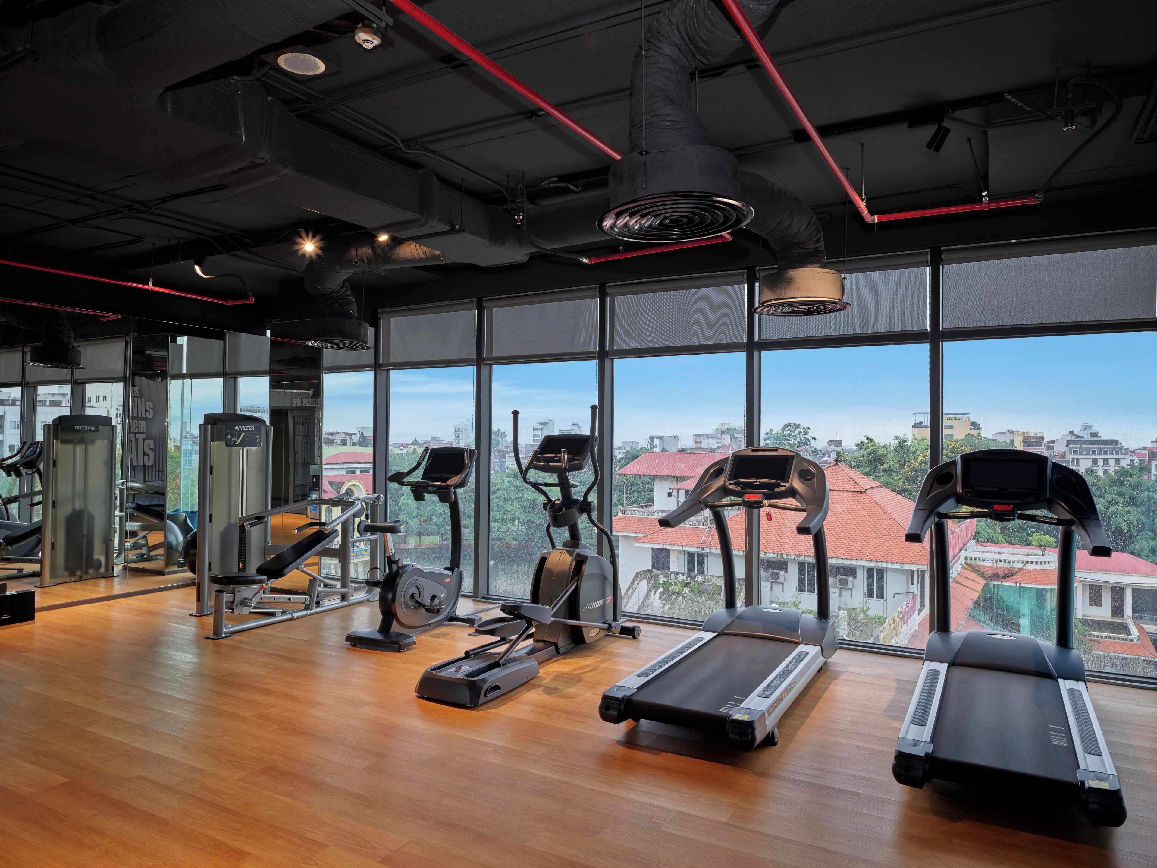 Une salle de sport équipée de tapis de course, de vélos elliptiques et d’appareils de musculation, avec de grandes fenêtres offrant une vue sur la ville.