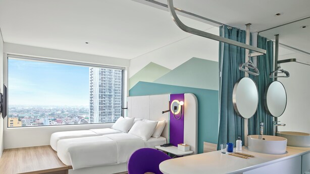 A modern hotel room with a large window, a bed, a desk, a chair, and a bathroom with a sink and mirror. - Tru by Hilton Bac Giang City Centre (Bac Giang)