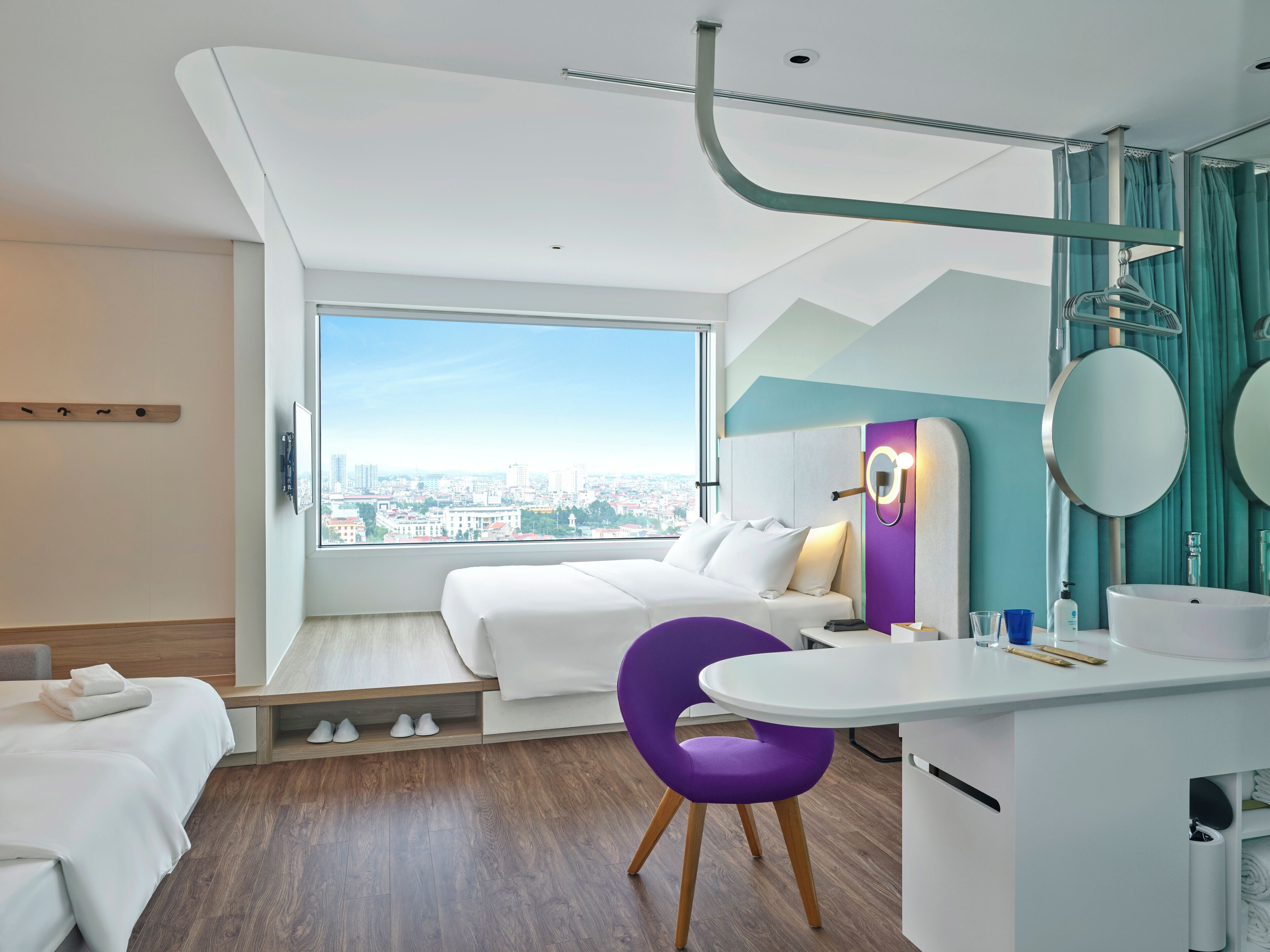 Una camera d'albergo moderna con un'ampia finestra, un letto, una scrivania con sedia e un bagno con specchio e lavandino.