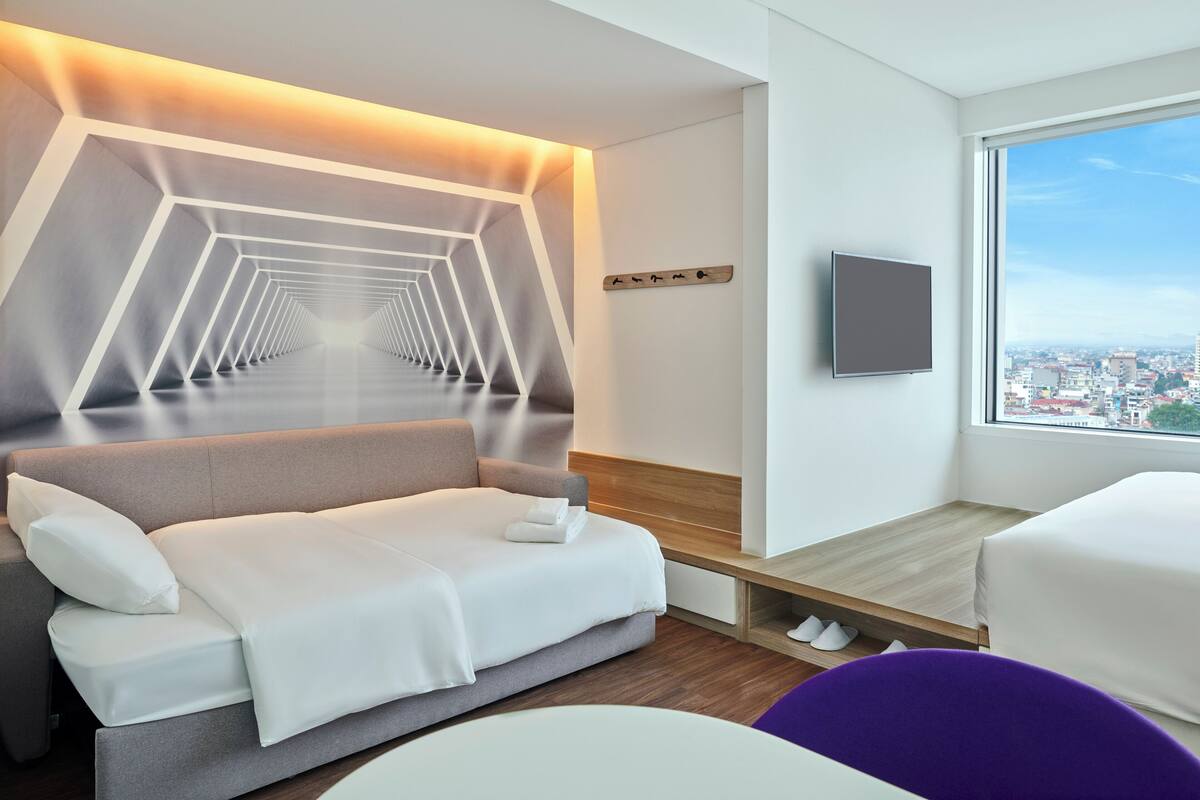 A modern hotel room with a sofa, a TV, a city view, and a unique geometric wall design.
