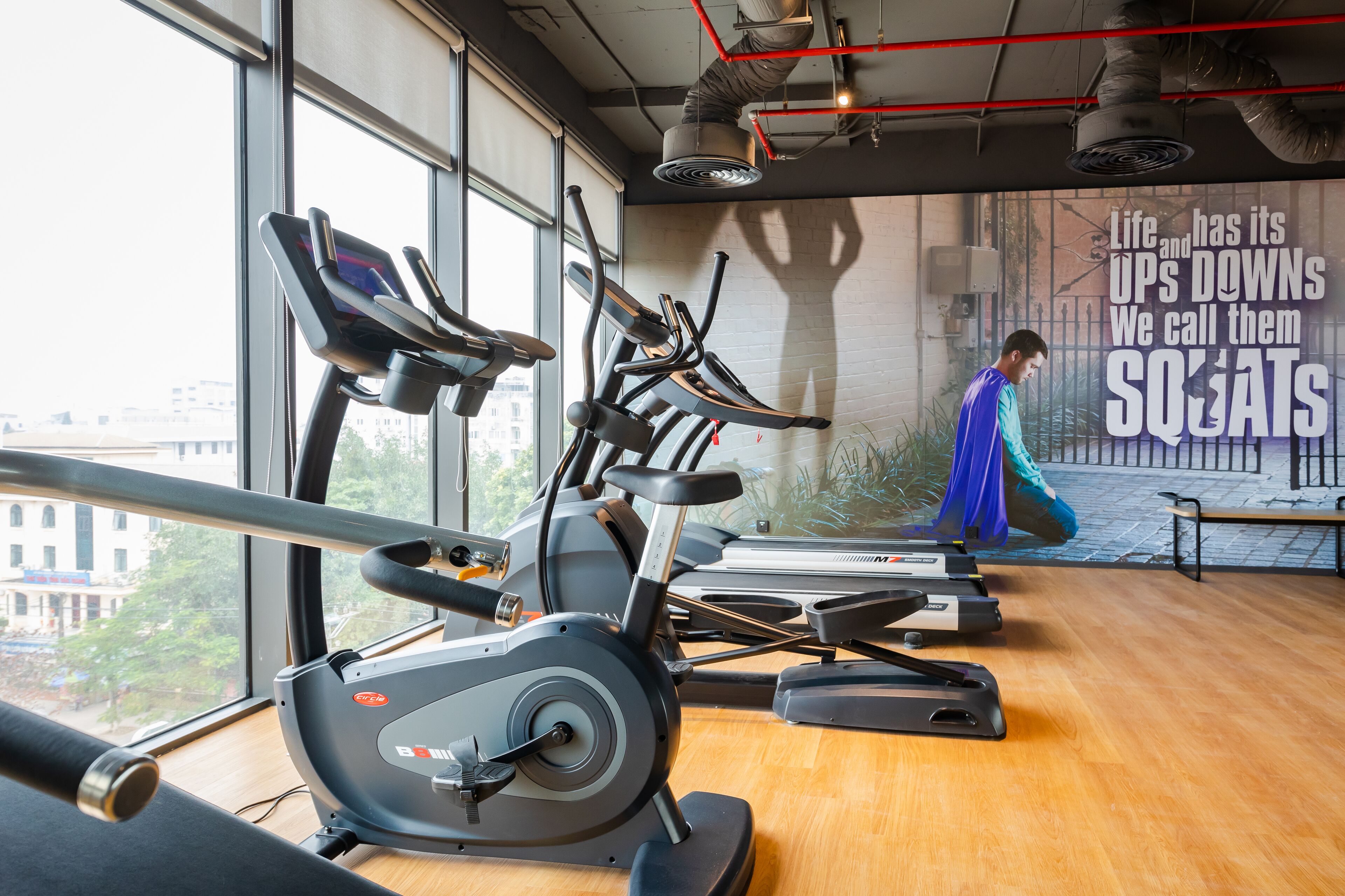 A gym with cardio machines, a mural with motivational text, and a view of buildings outside.