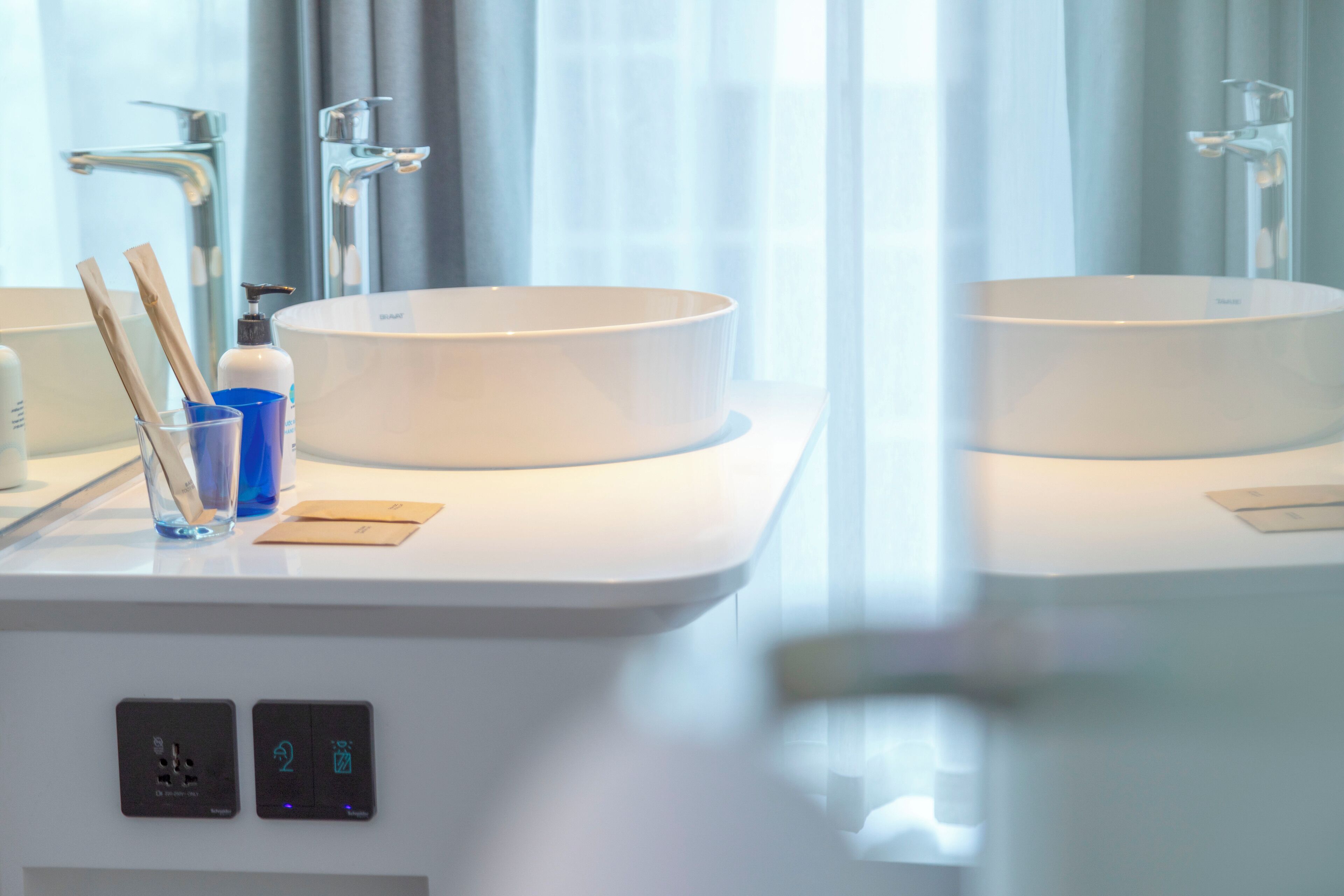 A modern bathroom with two round sinks, a mirror, and a window with curtains.