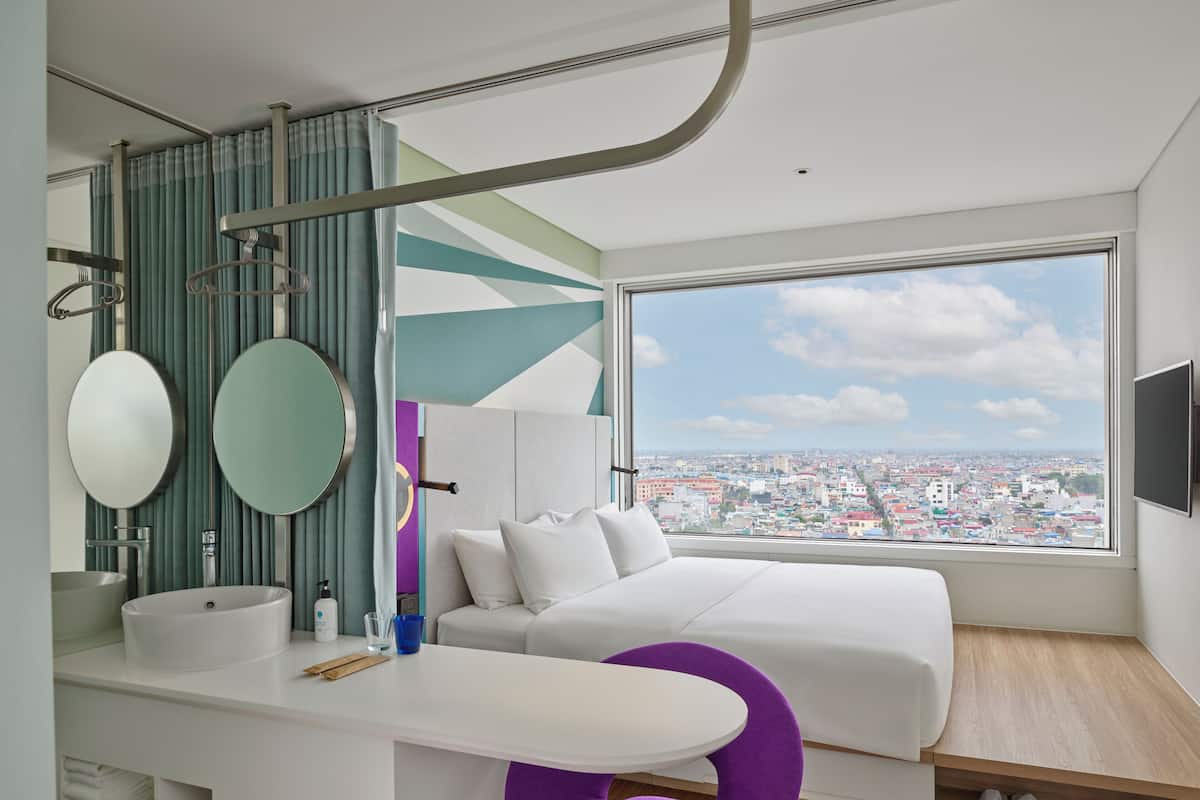 A modern bedroom with a large window, a bed, a vanity with a mirror, and a view of a cityscape.