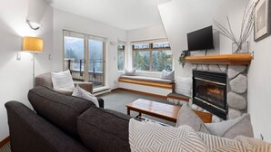 A modern living room with a fireplace, a sofa set, a television, and a large window overlooking a balcony. - 1BR | Lake Placid Lodge | Steps to Creekside Gondola | Shared Pool, Hot Tub, & Lounge (Whistler)
