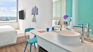 Una habitación de hotel moderna con un ventanal grande, una cama blanca, un lavamanos blanco y una silla color turquesa.
