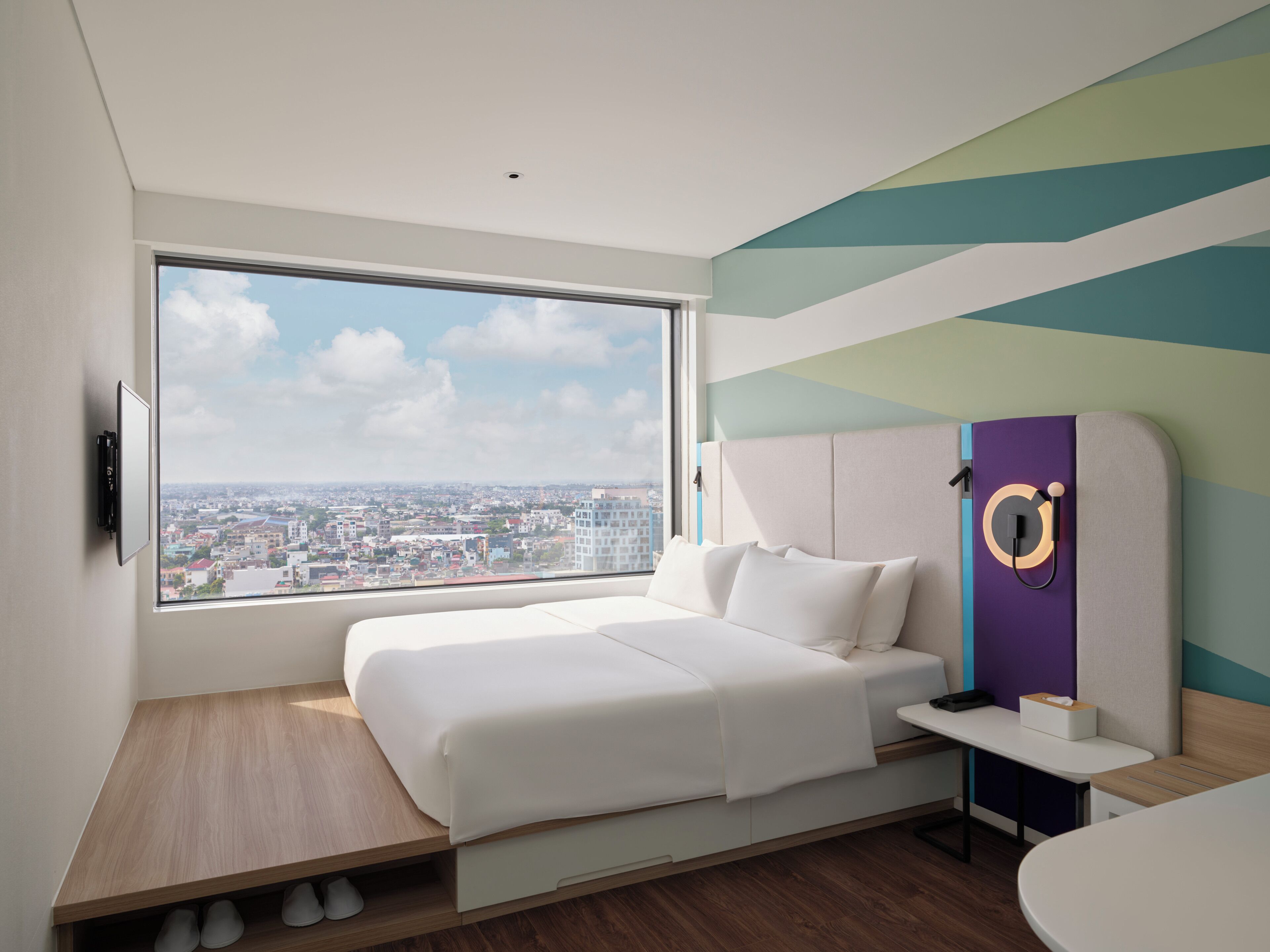 Una habitación de hotel moderna con un ventanal grande, una cama, una mesita de noche y un televisor montado en la pared.