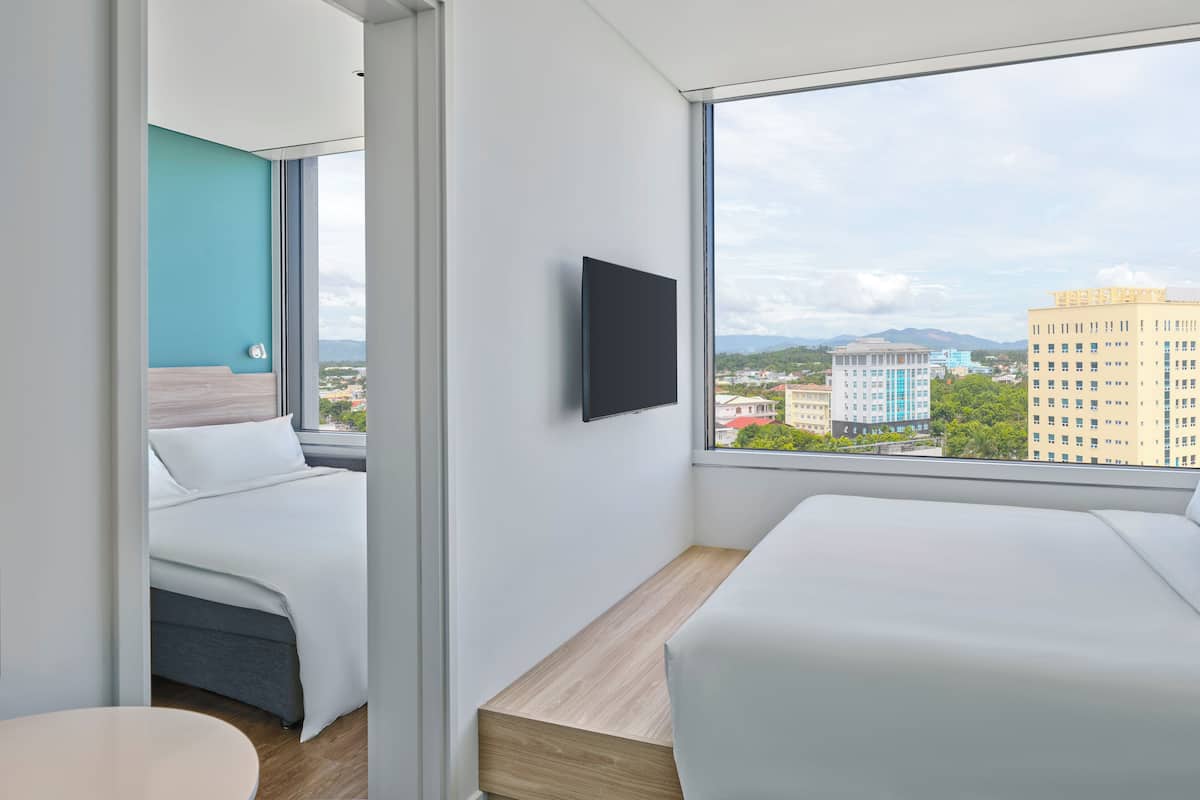 Ett modernt hotellrum med ett stort fönster som erbjuder utsikt över staden, en platt-TV som är monterad på väggen och en snyggt bäddad säng.