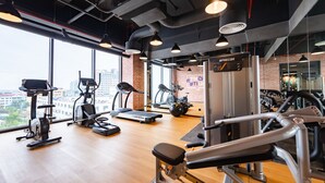 Un gimnasio con diversos equipos de ejercicio, amplias ventanas y una pared de ladrillo.