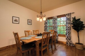 Un comedor con una mesa de madera, sillas, un candelabro, cuadros enmarcados, un ventanal con cortinas estampadas y una planta en maceta.