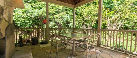 Une terrasse couverte avec un sol en pierre, une table et des chaises, et une vue sur un jardin luxuriant et verdoyant.