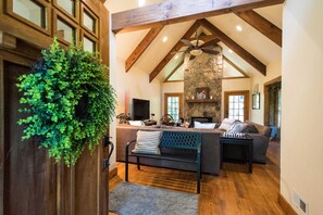 A spacious living room with a stone fireplace, wooden beams, a ceiling fan, and a large window.