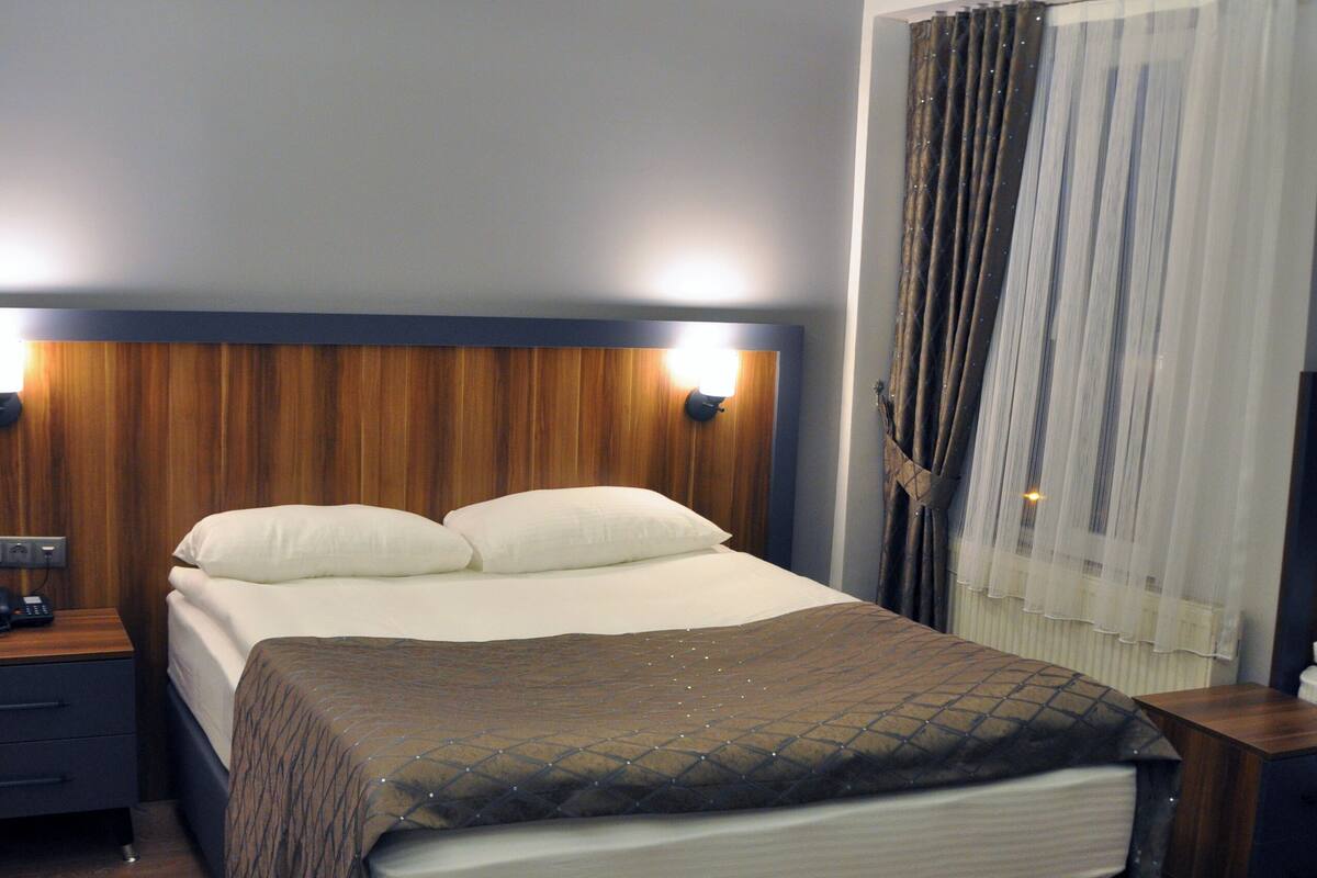 A hotel room with a large bed, wooden headboard, bedside tables, and a window with curtains.