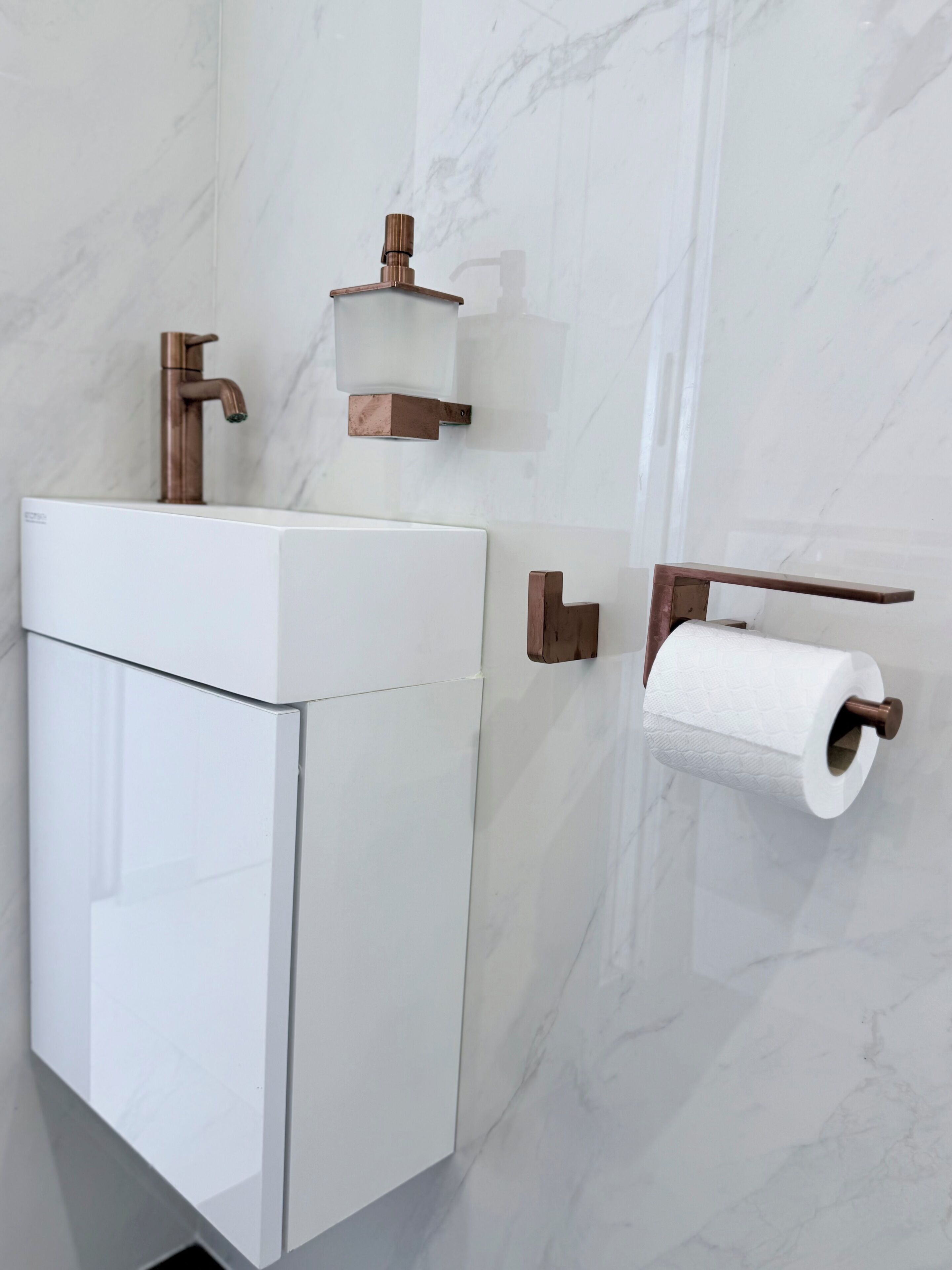 Een badkamer met een witte wasbak, een koperen kraan, een toiletpapierhouder en een marmeren wand.