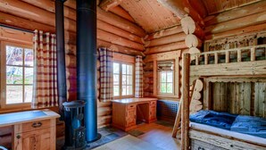 Interior de una cabaña de troncos con un fogón de leña en el centro, una cama litera y una pequeña zona de escritorio.