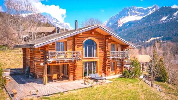 Una cabaña de madera con balcón, terraza y montañas al fondo.