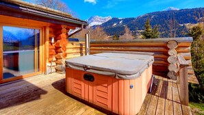 Una cabaña de madera con jacuzzi en una terraza, rodeada de montañas.