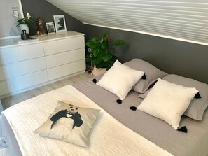 A bedroom with a bed, pillows, a bedside table, a plant, and a window with blinds.