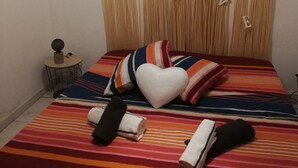 Ein Bett mit einem gestreiften Bettbezug, einem herzförmigen Kissen und aufgerollten Handtüchern. Auf einem kleinen runden Tisch steht ein sphärisches Objekt.