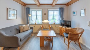 A cozy living room with a sofa, a table, and two chairs. There are windows with curtains, a television, and a radiator. - Guesthouse Am Badedeich, vacation apartment Sylt in Dagebüll for up to 4 people (Dagebüll)